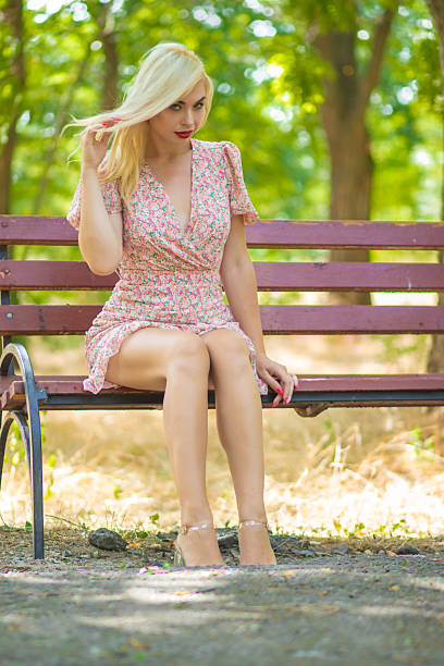 Blonde woman playing with her hair while sitting on a wooden bench in a sunny park. Stylish summer dress, red lipstick, natural bokeh.