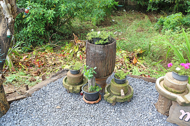 Creative garden decoration featuring vintage stone grinders and rustic wooden logs used as unique plant pots on a grey gravel floor.