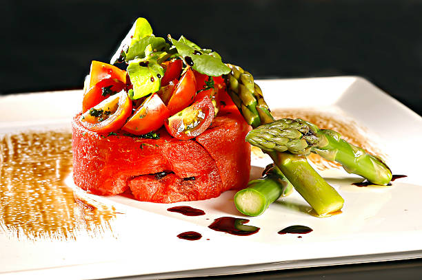 Close-up: Round slice of watermelon, chopped cherry tomatoes, chopped asparagus, arugula leaves, herbs and soy sauce, on a rectangular white plate, black background.