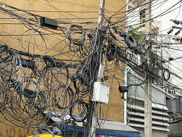 Stock photo showing electricity wires tangled and messy, in front of buildings in Bangkok, Thailand. The hazardous tangle of cables is due to the rapid growth in telecommunications with  providers choosing overhead installation since it is the cheaper alternative to the burying of cables as well as the sharing of poles with other utility power companies.