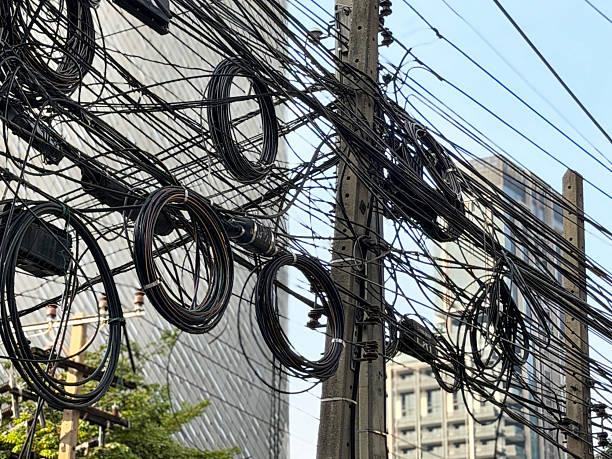 Stock photo showing electricity wires tangled and messy, in front of buildings in Bangkok, Thailand. The hazardous tangle of cables is due to the rapid growth in telecommunications with  providers choosing overhead installation since it is the cheaper alternative to the burying of cables as well as the sharing of poles with other utility power companies.
