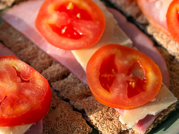 Stock photo shows nutritious snack featuring crisp rye crackers topped with savory ham, creamy cheese, and fresh tomato slices. Perfect for healthy eating, light meals, or balanced snacking, this wholesome combination highlights fresh ingredients, simple preparation, and appealing presentation