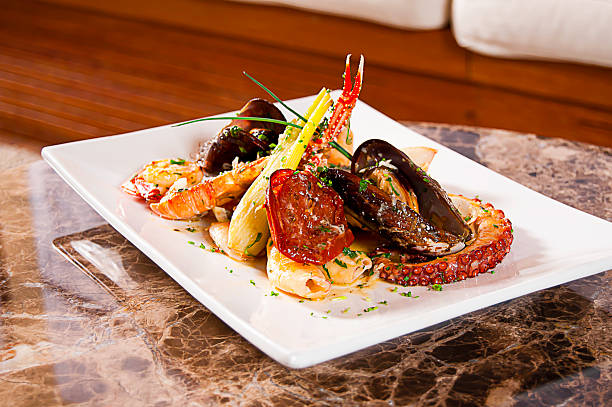 View: octopus, mussels in their shells, lobster, crab legs, fennel, sun-dried tomatoes, fresh herbs, shallots, sauce, rectangular white porcelain plate, on a stone table, wood background.