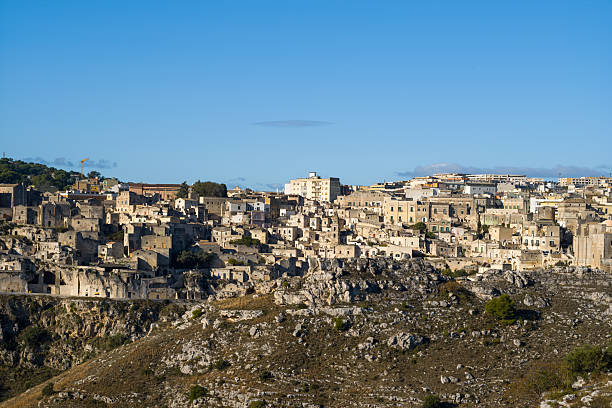 Expansive view of Matera, Italy, with ancient stone houses densely packed on rugged hillsides, bathed in crisp sunlight and framed by a vivid blue sky.
