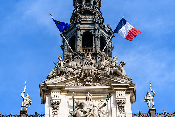 Elaborate stone sculptures of allegorical figures and classical statues adorn the ornate rooftop of Hotel de Ville in Paris, with French and European Union flags waving against a clear blue sky.
