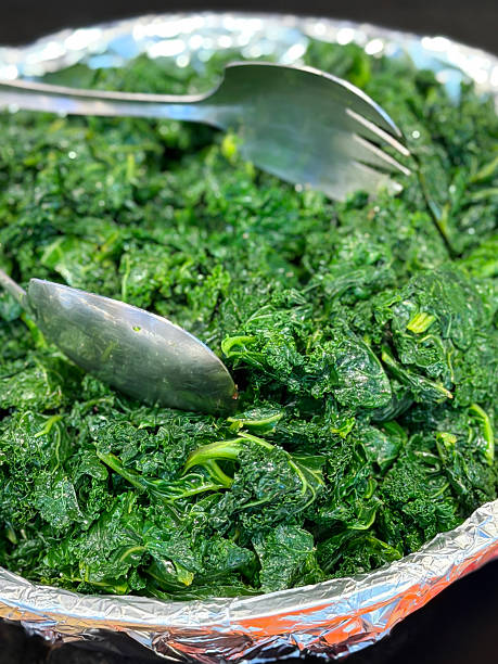 Full frame image of aluminium foil lined bowl with pile of steamed kale under heat lamp, stainless steel serving tongs, healthy side dish option, restaurant buffet service, elevated view