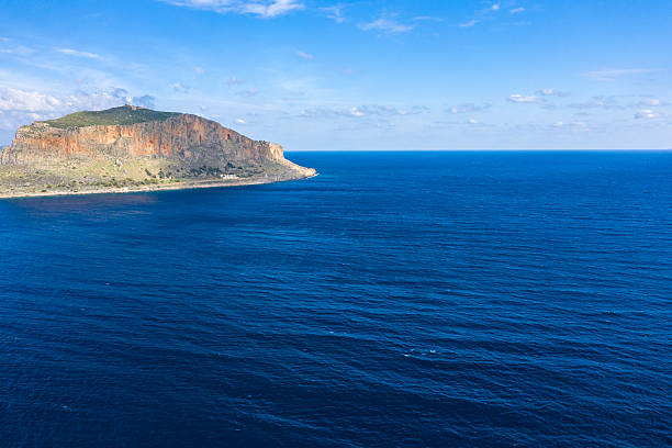 Expansive view of deep blue Mediterranean waters stretching toward the rugged cliffs of Monemvasia, Greece. Bright sunlight enhances the vivid colors and textures beneath a clear sky with scattered clouds.