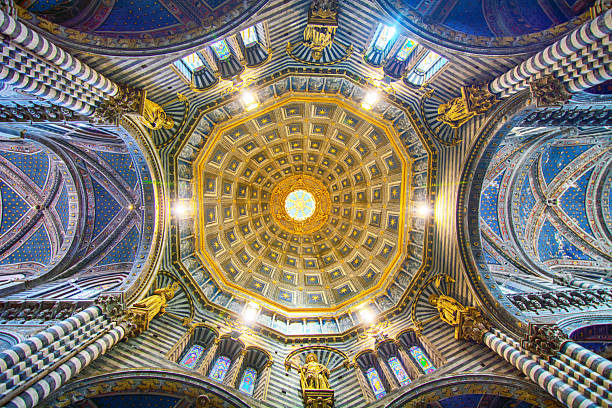 This image captures the breathtaking interior of a grand cathedral, likely the Siena Cathedral (Duomo di Siena) given its distinctive architectural elements. The most striking feature is the ceiling, a magnificent dome adorned with intricate gold detailing and geometric patterns, radiating outwards from a central point. Light streams in from windows near the top, illuminating the ornate design.Below the dome, the architecture is characterized by alternating horizontal bands of dark gray or black and white marble on the massive columns and arches, creating a visually striking and rhythmic pattern that extends throughout the interior. Ornate carvings, statues, and decorative elements embellish the arches and walls, showcasing skilled craftsmanship.Stained glass windows with vibrant colors are visible, filtering light into the space. In the lower portion of the image, an elaborately decorated altar or chapel area is visible, featuring gilded details and religious artwork. People can be seen as small figures in the lower part of the frame, giving a sense of the immense scale of the cathedral. The overall atmosphere is one of awe-inspiring grandeur, artistic richness, and historical significance. The warm lighting from within contrasts with the cooler tones of the marble, creating a dramatic and reverent ambiance.