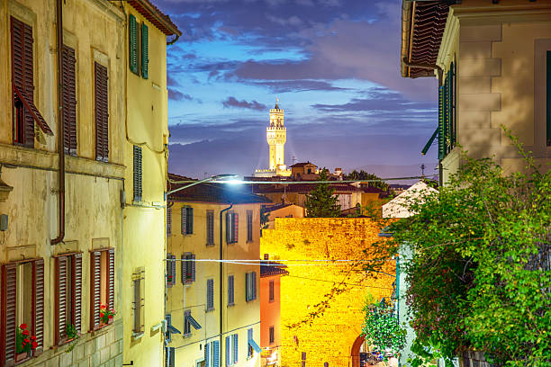 A twilight view of Florence with narrow historic streets leading toward the illuminated tower of Palazzo Vecchio rising above the rooftops