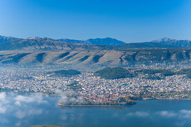 A wide panoramic scene captures the city of Ioannina nestled beside Lake Ioannina, with the dramatic ridges of the Pindus mountains rising in the background. Low clouds drift above the calm water, highlighting the natural textures and urban landscape of Epirus, Greece.