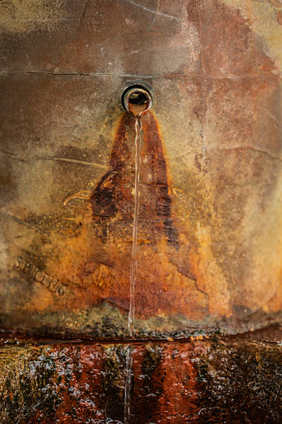 Close-up of water dripping from a small opening in a rust-stained stone fountain, highlighting weathered texture, mineral deposits, and natural aging.