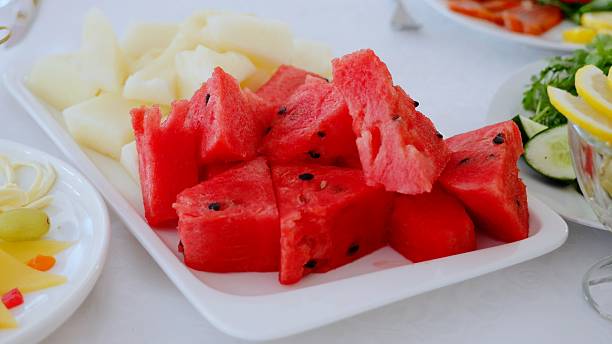 Freshly cut watermelon and melon are served on a white rectangular plate, part of a larger buffet spread including cheese, grapes, smoked salmon, cucumbers, and lemon