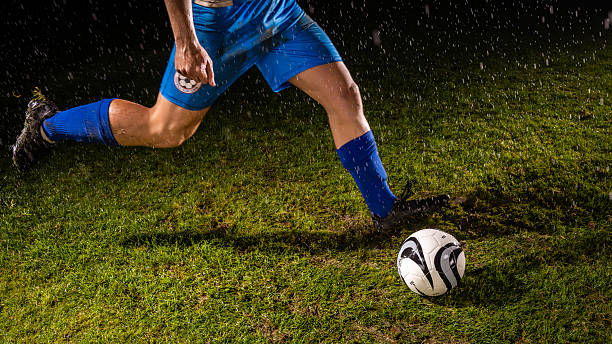 A soccer player in blue shorts and socks intensely controls a white and black ball on a grassy field. Raindrops highlight determination and skill during practice, capturing sports training and dedication.