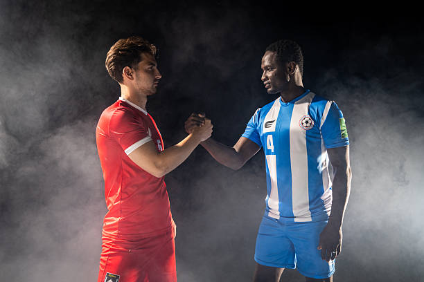 Two soccer players, one in a red jersey and the other in a blue and white striped jersey, shake hands against a smoky backdrop, showcasing sportsmanship and camaraderie.