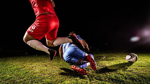 Soccer players in action on a grassy field at night. One in red runs, the other in blue slides to tackle. The ball is nearby, capturing a dynamic moment of competition and skill in a sports training setting.