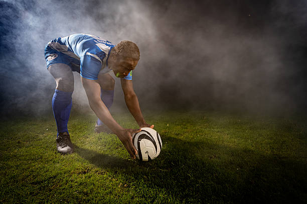 A focused soccer player in a blue uniform bends to place a ball on a grassy field, surrounded by a misty atmosphere. The scene captures the intensity and preparation of a soccer practice session, highlighting the athlete's dedication and the sport's dynamic environment.