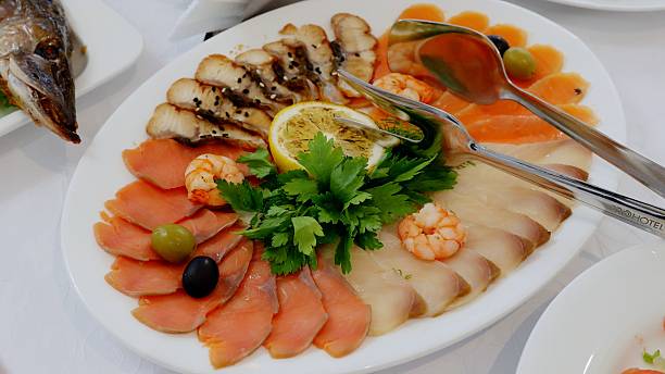 Beautifully arranged platter of various fish delicacies, including smoked fish, salmon slices, shrimp, lemon, and olives, is presented on a white tablecloth, ready to be enjoyed