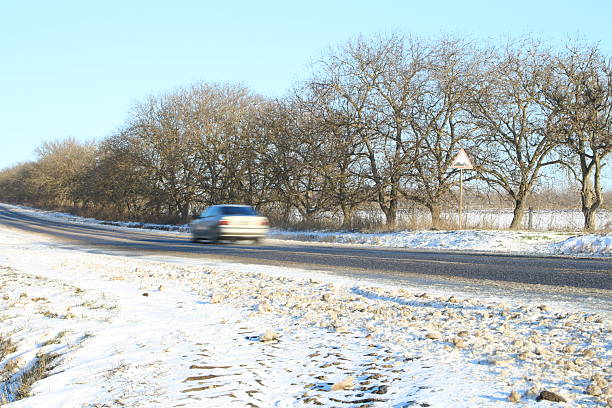 A car driving on a road with snow on the ground and trees. Tree. Outdoor. Nature. Shore. Sandy