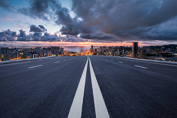 Dramatic sunset sky hangs over an empty asphalt road leading toward a modern coastal city skyline.