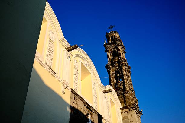 A yellow church facade catches strong sunlight beside a carved stone bell tower under a deep blue sky.