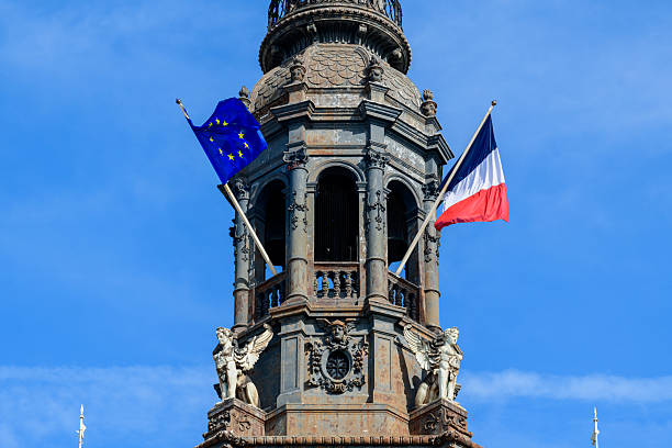Decorative stone bell tower of Hotel de Ville in Paris displays the French and European Union flags, flanked by sculpted angel figures and highlighted against a bright blue sky.