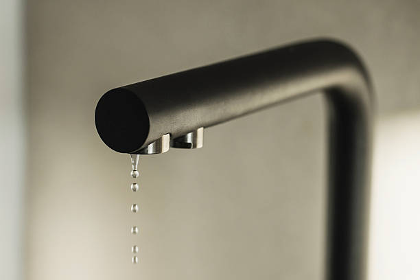 A modern metal kitchen water tap with a drop of water hanging down from it, isolated on the black background. Selective focus on the drop of water.