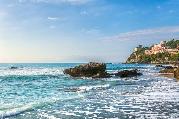 View of the rocky Quercetano bay beach in Castiglioncello. Coastal landscape with blue sea waves and historic liberty hotels on the cliff, Tuscany, Italy