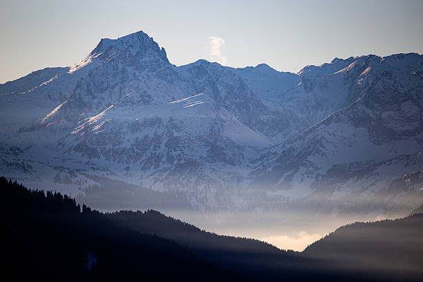 Snow covered mountain peaks rising above layers of mist. Peaceful winter mountain landscape with soft light and clear sky.