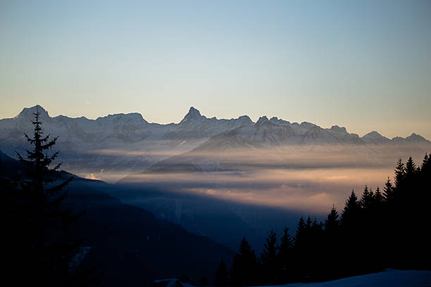 Scenic winter landscape of alpine mountains with morning mist, snow peaks and warm sunrise light.
