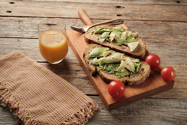 Two slices of bread covered with avocado are placed on a wooden board. There are cherry tomatoes nearby and a glass of orange juice accompanies the meal.