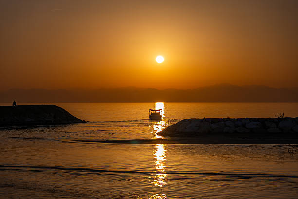 Silhouette of a small boat sailing through the golden reflection of the setting sun between two piers at the ocean.
