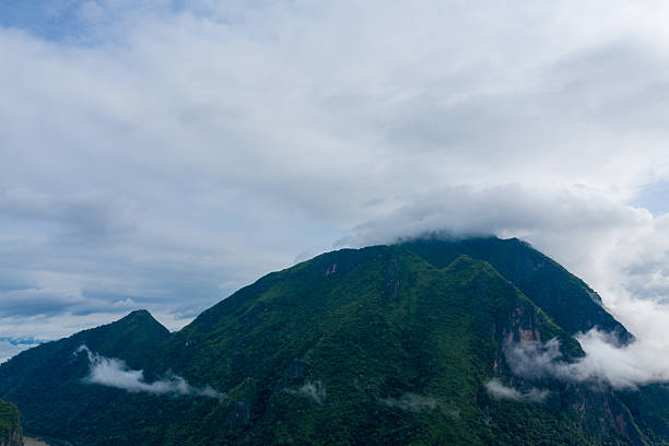 Low clouds and mist move across steep, forested mountains near Nong Khiaw, Laos. The lush green slopes and dramatic sky create a tranquil natural landscape under soft daylight.
