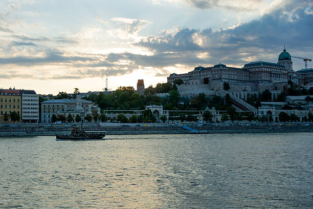 Budapest, Hungary - September 2025: The historic Buda Castle (Budavari Palota) complex stands on the hill overlooking the Danube river. In the foreground, a traditional paddle steamer tourist boat sails on the river. The Varkert Bazar and the dome of the Royal Palace are visible under a cloudy sky with evening light.