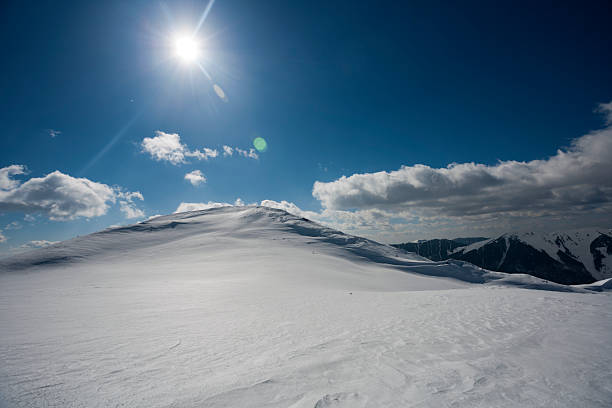 A landscape shot of a massive, snow-blanketed mountain range beneath a brilliant, deep blue sky. The foreground highlights the unique wind-blown textures and ripples in the snow, leading the viewer's eye toward several sharp, sunlit peaks. The image evokes a sense of cold, quiet majesty and the vastness of the alpine wilderness, perfect for travel, adventure, and nature-themed projects.