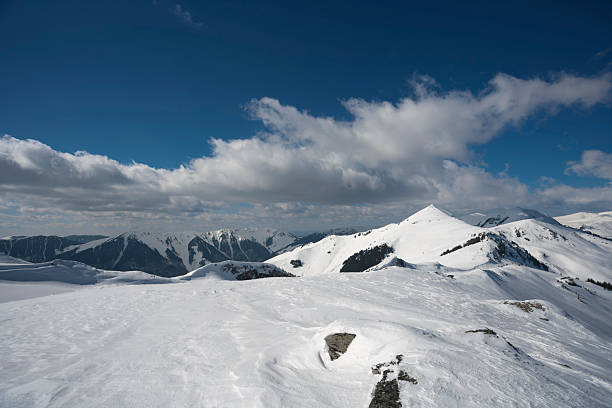 A landscape shot of a massive, snow-blanketed mountain range beneath a brilliant, deep blue sky. The foreground highlights the unique wind-blown textures and ripples in the snow, leading the viewer's eye toward several sharp, sunlit peaks. The image evokes a sense of cold, quiet majesty and the vastness of the alpine wilderness, perfect for travel, adventure, and nature-themed projects.