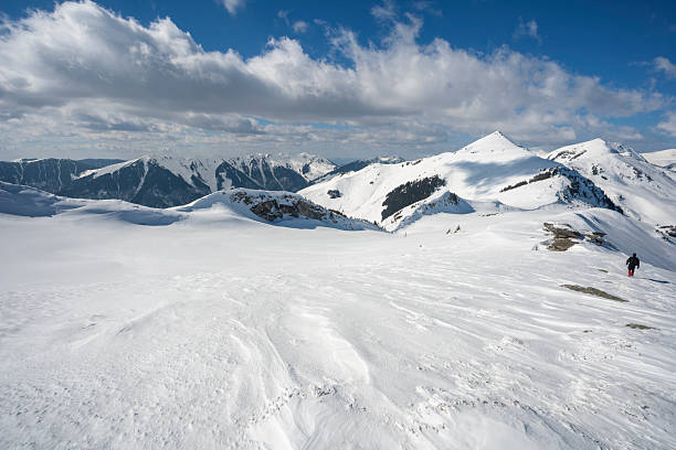 A landscape shot of a massive, snow-blanketed mountain range beneath a brilliant, deep blue sky. The foreground highlights the unique wind-blown textures and ripples in the snow, leading the viewer's eye toward several sharp, sunlit peaks. The image evokes a sense of cold, quiet majesty and the vastness of the alpine wilderness, perfect for travel, adventure, and nature-themed projects.