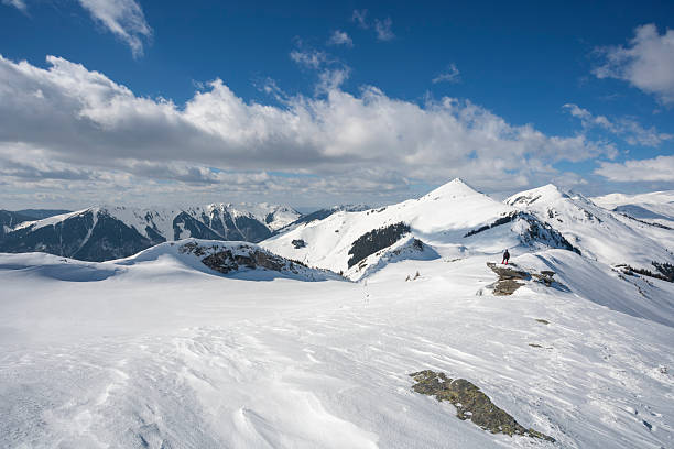 A landscape shot of a massive, snow-blanketed mountain range beneath a brilliant, deep blue sky. The foreground highlights the unique wind-blown textures and ripples in the snow, leading the viewer's eye toward several sharp, sunlit peaks. The image evokes a sense of cold, quiet majesty and the vastness of the alpine wilderness, perfect for travel, adventure, and nature-themed projects.