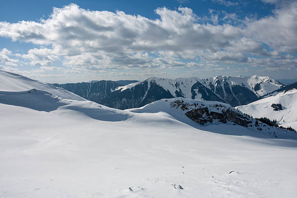 A landscape shot of a massive, snow-blanketed mountain range beneath a brilliant, deep blue sky. The foreground highlights the unique wind-blown textures and ripples in the snow, leading the viewer's eye toward several sharp, sunlit peaks. The image evokes a sense of cold, quiet majesty and the vastness of the alpine wilderness, perfect for travel, adventure, and nature-themed projects.