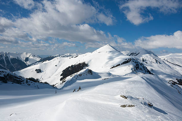 A landscape shot of a massive, snow-blanketed mountain range beneath a brilliant, deep blue sky. The foreground highlights the unique wind-blown textures and ripples in the snow, leading the viewer's eye toward several sharp, sunlit peaks. The image evokes a sense of cold, quiet majesty and the vastness of the alpine wilderness, perfect for travel, adventure, and nature-themed projects.