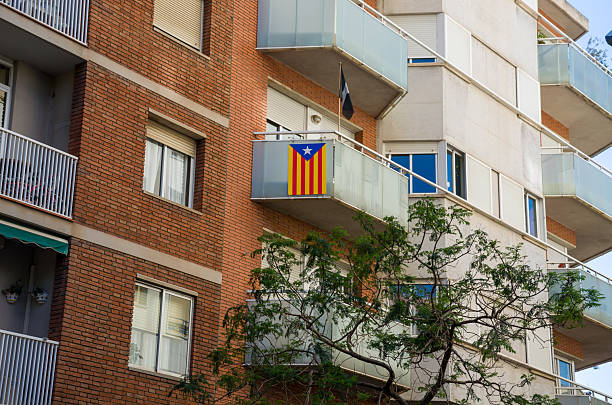 Modern apartment building with brick and concrete facade featuring a balcony displaying the Catalan independence flag and a tree in front.