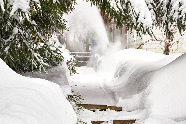 Man clearing a snow-covered garden path with a snow blower after heavy snowfall. Winter scene with deep snowbanks, evergreen trees, cold weather, seasonal work, and residential outdoor maintenance