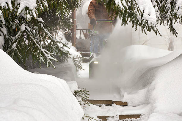 Man clearing a snow-covered garden path with a snow blower after heavy snowfall. Winter scene with deep snowbanks, evergreen trees, cold weather, seasonal work, and residential outdoor maintenance