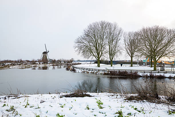 Tranquil winter scene featuring a traditional windmill beside a frozen lake, snow-covered ground, and leafless trees, Haastrecht, Netherlands.