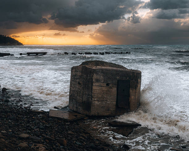 A dramatic view of an abandoned concrete military bunker (pillbox) situated on a rocky coastline. Rough sea waves crash against the old structure, while golden sunset light breaks through dark storm clouds in the background.