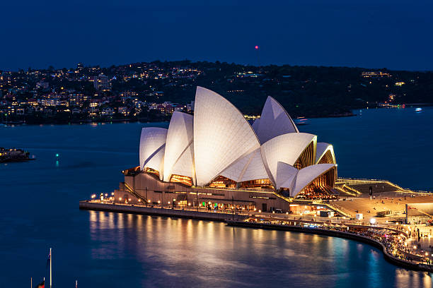 Sydney, Australia - The Sydney Opera House illuminated at dusk, with the light reflecting in the water of Sydney Harbour.