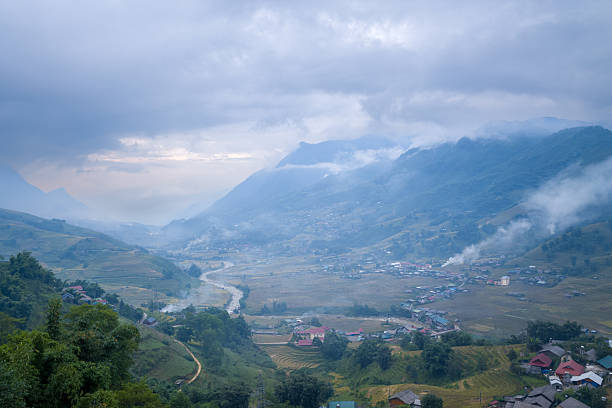 A winding river flows through a broad mountain valley in Sapa, Vietnam, with scattered villages, terraced fields, and drifting morning mist under dramatic cloudy skies.