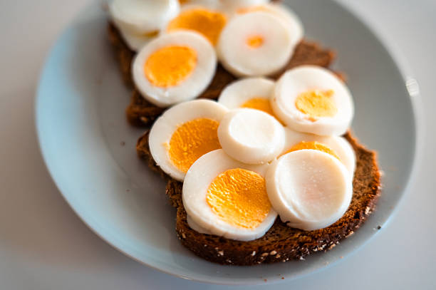 Top view of open-faced sandwiches topped with sliced hard-boiled eggs on whole grain bread, served with fresh cucumber sticks on a white plate. Minimalist food styling with natural light and clean background, representing healthy breakfast, simple home cooking, balanced nutrition and everyday lifestyle concepts.