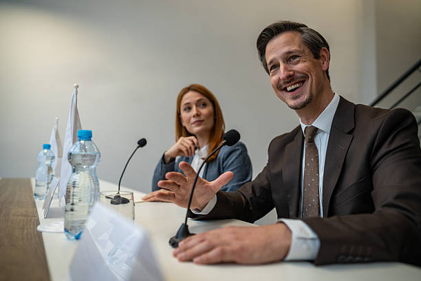 Two diplomats in suits sit at a conference table with microphones, exchanging ideas and smiling during a political summit on international relations and governance policy