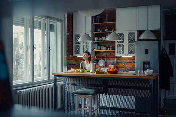 Happy woman laughing, looking through window, preparing healthy fresh smoothie in a modern kitchen. Domestic lifestyle and wellness concept