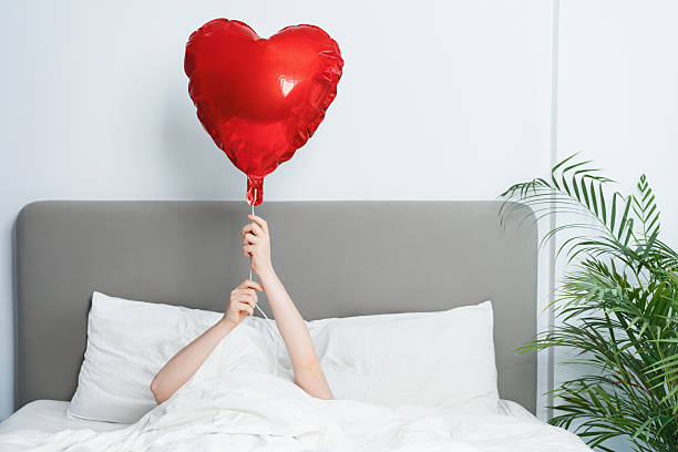 A person lies alone in a white bed holding a red heart balloon, expressing loneliness and unmet love on Valentine day indoors